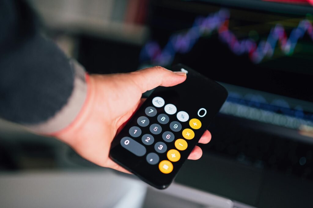 pexels-photo-20552602-20552602 Close-up of a hand holding a smartphone calculator with financial charts on a screen in the background.