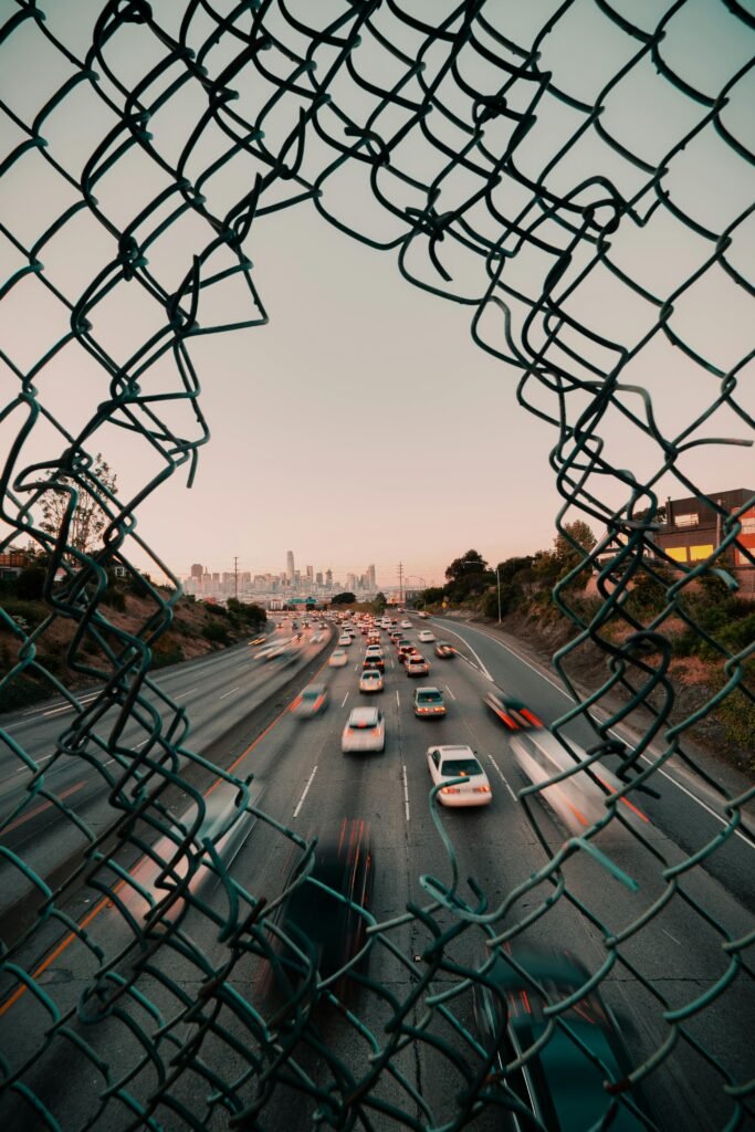 pexels-photo-3849168-3849168 Traffic flows on a highway framed by a broken fence with San Francisco cityscape at sunset.