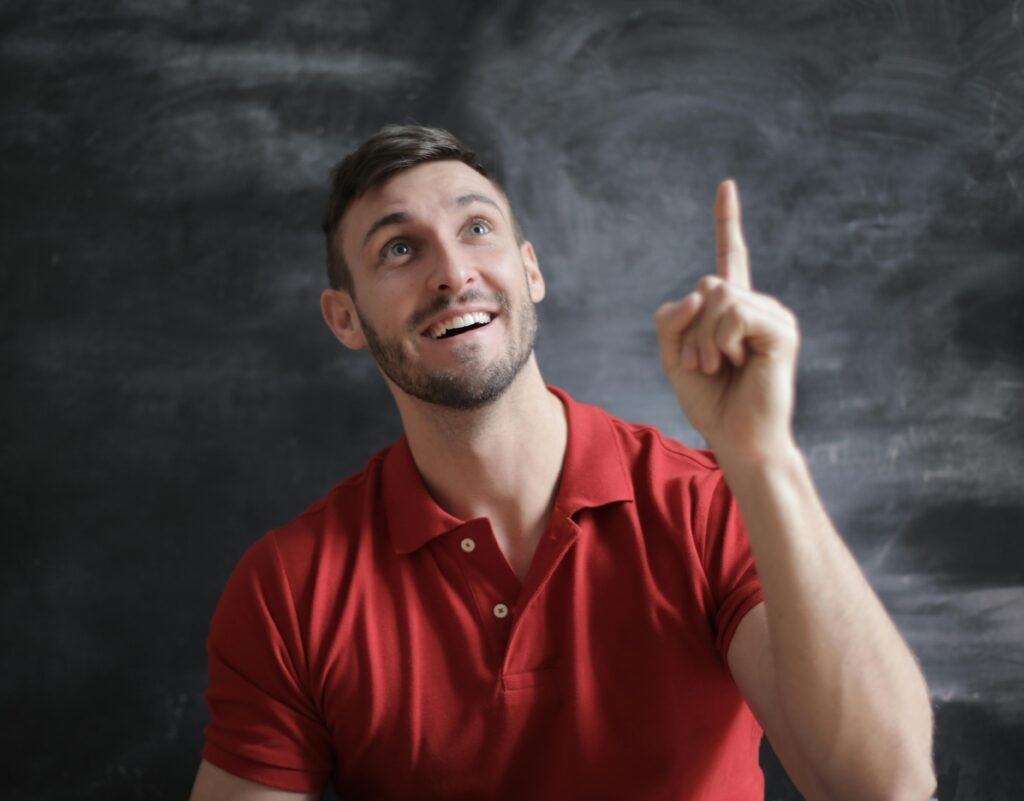 pexels-photo-3779432-3779432 Happy young man in a red polo shirt smiling and pointing upwards in front of a blackboard.