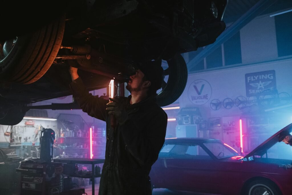 pexels-photo-4488654-4488654 A mechanic inspects a car's undercarriage in a vibrant, neon-lit garage setting, highlighting automotive repair work.