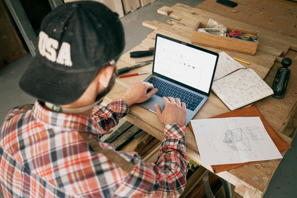 A woodworker in a workshop uses a laptop for furniture design planning, project management, and building passive income streams.